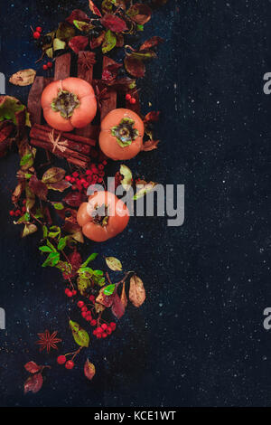 Ripe persimmons on a dark wooden background with autumn leaves, berries and cinnamon. Flat lay with copy space. Fruit in dark food photography.