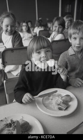 1970s, historical, picture shows a young girl eating her school dinner (lunch) nicely with a knife and fork sitting next to a young boy with other excited children looking on, Langbourne Primary School, Dulwich, SE21, London, England, UK. Stock Photo