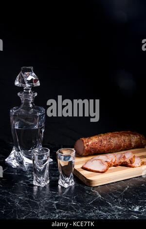 Bottle and shot glasses with vodka. Slices of smoked meat or ham on wooden cutting board. Composition on black marble background. Stock Photo