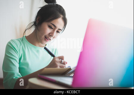 Young attractive Asian woman freelancer writing on notebook while working with laptop computer. Work at home and online learning concept Stock Photo