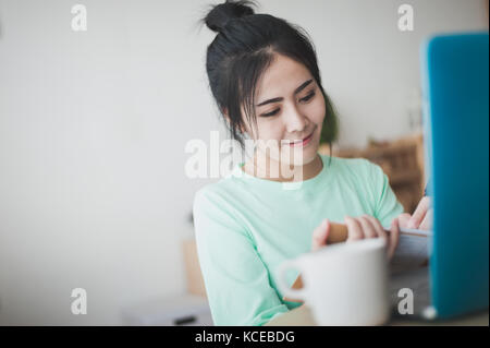 Young attractive Asian woman freelancer writing on notebook by left hand while working with laptop computer. Work at home and online learning concept Stock Photo