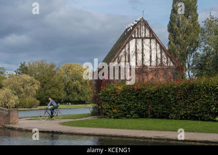 Sunny, autumnal view of a man on a bike cycling by the lake & lych-gate in beautiful, peaceful, municipal Rowntree Memorial Park, York, England, UK. Stock Photo