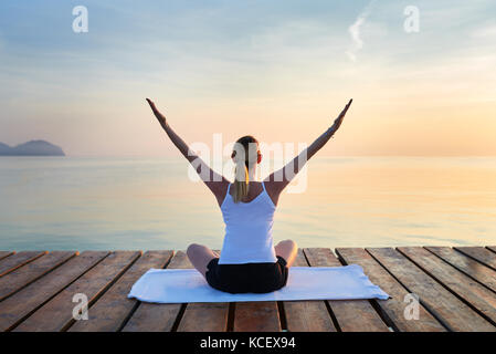 Rear view of young woman sat on towel practising yoga by sea at sunset Stock Photo