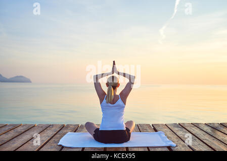 Rear view of young woman sat on towel practising yoga by sea at sunset Stock Photo