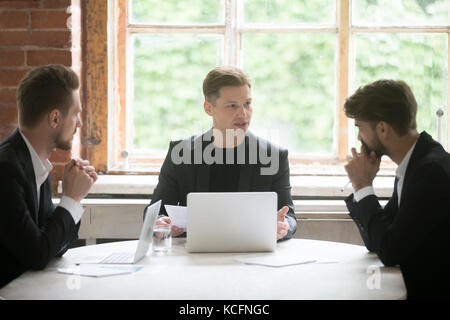 Young serious boss in front of laptop consulting two male employees during briefing meeting. Executive employee talking about company goals to coworke Stock Photo