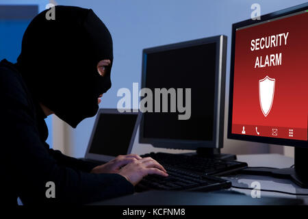 Burglar Wearing A Balaclava Breaking Security Alarm On Computer In Office Stock Photo