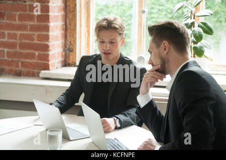 Two male executives looking at laptop screen during business meeting. Businessmen discuss corporate goals in front of laptops at workplace in office.  Stock Photo