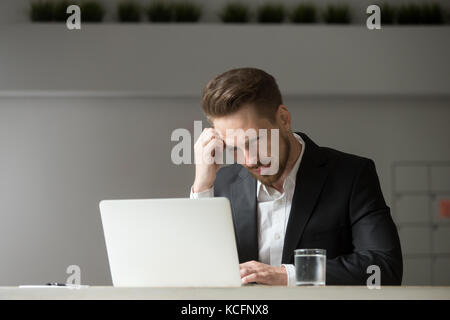 Perplexed, puzzled handsome businessman looking at laptop screen at workplace in office. Young CEO or project manager is not sure whether he should ag Stock Photo