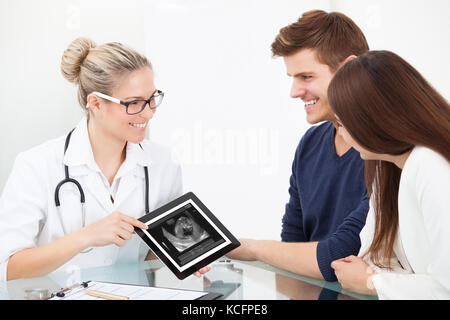 Female Doctor Showing Ultrasound Scan Of Baby On Digital Tablet To Happy Couple At Clinic Stock Photo