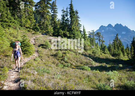 A young woman hiking with backpack along trail to 5040 Peak in the Alberni-Clayoquot region on Vancouver Island, BC, Canada. Triple Peak can be seen to the right in the background. Stock Photo