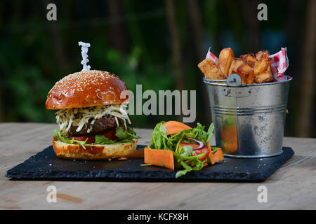 Cheeseburger with tomato, rocket, salad, coleslaw in a brioche bun and a bucket of chunky chips, fries. Stock Photo