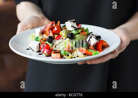 The young female waiter holding in her hands the plate with the Greek salad Stock Photo