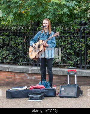 A smiling young woman busker, singing and playing acoustic guitar, with microphone and portable amplifier, on the South Bank, London, England, UK Stock Photo