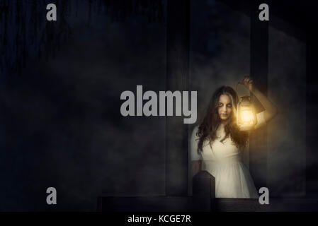 Spooky image of a scary woman with dark eyes and appearance of a witch, in a white dress, holding a lit lantern, in a dark night atmosphere. Stock Photo