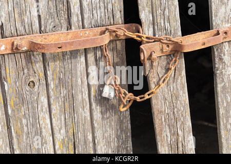 old rusty lock and a metal chain, bolted on a broken wooden door. Photo close-up. In the door panels there are a number of damages, and several bars h Stock Photo