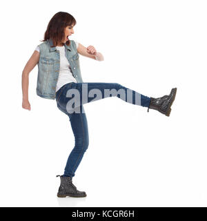 Young woman in jeans vest and black boots is shouting and kicking. Side view. Full length studio shot isolated on white. Stock Photo
