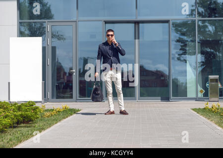 Young official man expert calling via cell telephone while standing outdoors near modern glass building, young male employer talking on mobile phone Stock Photo