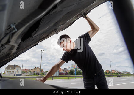 Closeup portrait, young man having trouble with his broken auto, opening hood trying to fix engine, isolated green trees outside background. Stock Photo