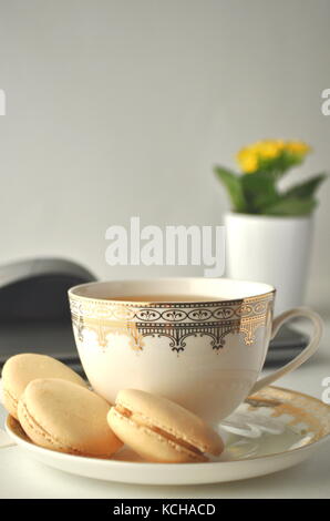 Cup of tea with French macaroons, laptop computer, mouse, pen and yellow plant on a table. Work space of a freelancer. Stock Photo