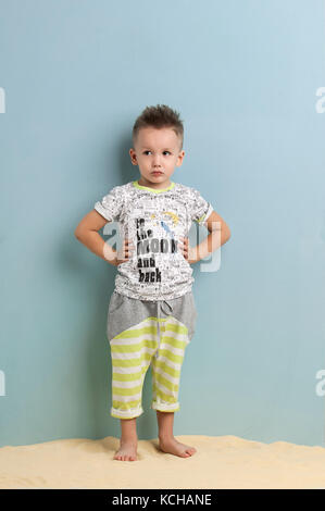 little boy in shorts and t-shirt standing on the sand on a light blue background Stock Photo