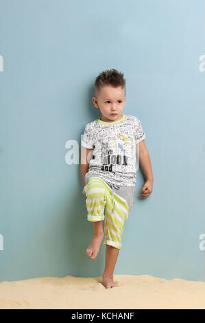 little boy in shorts and t-shirt standing on the sand on a light blue background Stock Photo