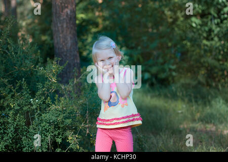 Little beautiful blonde girl standing in the forest. She girl put her face in her hands and bowed her head, she laughs Stock Photo