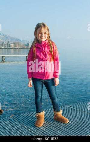 Outdoor portrait of cute little girl wearing pink jacket, resting next to lake Stock Photo