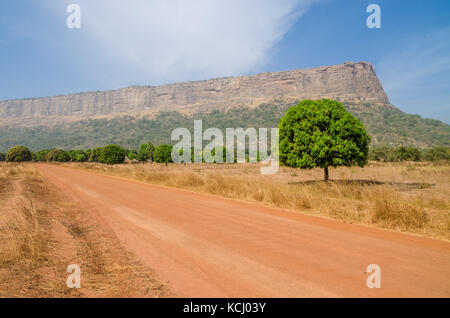 Red dirt and gravel road, single trees and large flat topped mountain in Fouta Djalon region, Guinea, West Africa Stock Photo