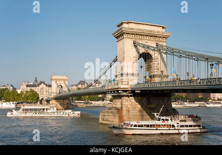 A view of the Széchenyi Chain Bridge in Budapest with a day trip river cruise boat passing underneath on a sunny day with blue sky. Stock Photo