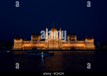 An evening nighttime view of the Hungarian Parliment Building on the Danube river in Budapest with a tourist river boat cruise vessel going past. Stock Photo