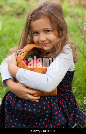 Portrait Of Cute Little Six-Year Girl Sits And Hugging Wicker Basket With Red Ripe Apples In Garden In Summer. Close-up. Stock Photo