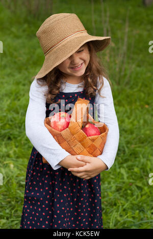 Happy Joyful Little Six Year Old Girl In Wicker Hat Hold In Her Hands Wicker Basket With Red Ripe  Apples In Garden Close Up. Little Girl With Red App Stock Photo