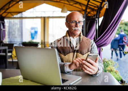 Old senior businessman in suit and tie with laptop computer and smartphone, sitting in city cafe house Stock Photo