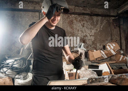 A young man in a black T-shirt, a black welding mask smiles and brews a metal welding machine in a dark workshop, in the background of equipment, hose Stock Photo