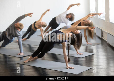 Group of young sporty people practicing yoga lesson with instructor, stretching in Bending Side Plank exercise, Vasisthasana pose, working out, indoor Stock Photo