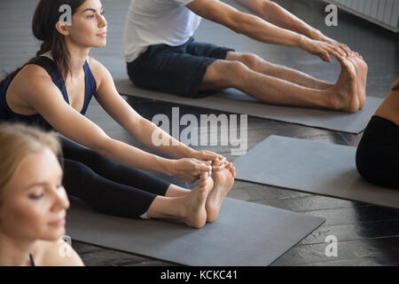 Group of young sporty people practicing yoga lesson with instructor concept, sitting in paschimottanasana exercise, Seated forward bend pose, friends Stock Photo
