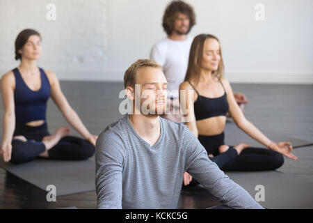 Group of young people, sporty students practicing yoga lesson with instructor, sitting and meditating with closed eyes in Padmasana exercise, Lotus po Stock Photo