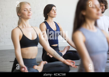 Group of sporty people, young students practicing yoga lesson with instructor, sitting and meditating with closed eyes in Padmasana exercise, Lotus po Stock Photo