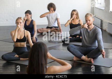 Group of young sporty people practicing yoga lesson with instructor, making nadi shodhana pranayama exercise sitting in Sukhasana pose, friends workin Stock Photo