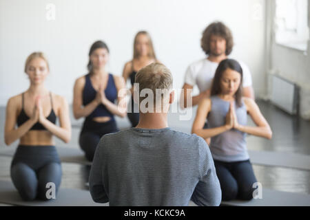 Group of young sporty people practicing yoga lesson with instructor, sitting in vajrasana exercise, seiza pose, working out indoor, studio, rear view  Stock Photo