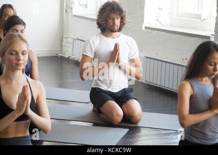 Group of young sporty people practicing yoga lesson with instructor, sitting in vajrasana exercise, seiza pose, working out, indoor full length, studi Stock Photo