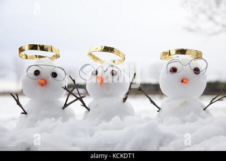 Three cute snowman angles with golden halos and reading glasses and their wooden twig arms in the air. Snow fall on the ground at winter Stock Photo