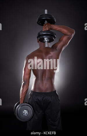 Rear View Of An African Man Exercising With Dumbbells On Black Background Stock Photo