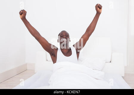 Young Happy Man Raising Arm While Sitting On Bed Stock Photo