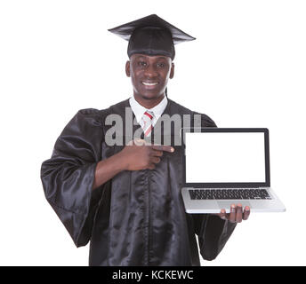 Male Student In Graduate Robe Pointing Towards Laptop Over White Background Stock Photo