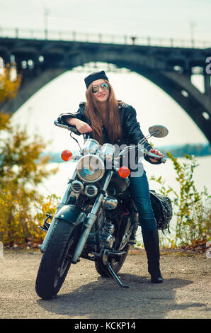 Smiling young woman in a leather jacket and glasses on a motorcycle on a urban autumn background. Stock Photo