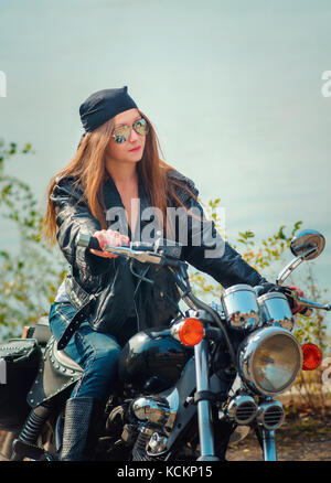 Beautiful young woman in a leather jacket  and blue jeans in a glasses on a motorcycle on a urban autumn background. Stock Photo