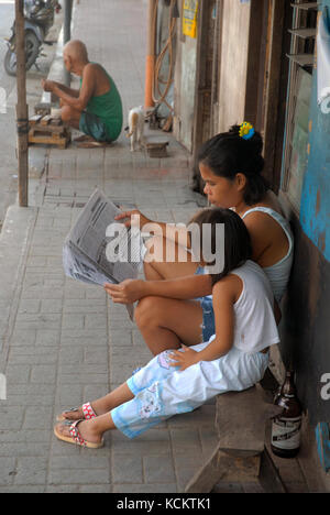 Mother reading a newspaper whilst sat on pavement with her daughter, Cebu, Philippines. Stock Photo