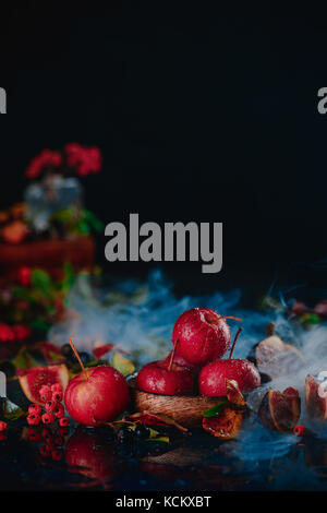 Tiny red apples on a wooden plate in an autumn still life with leaves and smoke. Dark food photography with copy space. Stock Photo