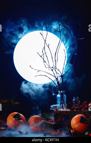 Tree branches silhouette in full moon. Spooky Halloween concept with autumn fruits, berries, magical books and smoke. Magical equipment still life wit Stock Photo